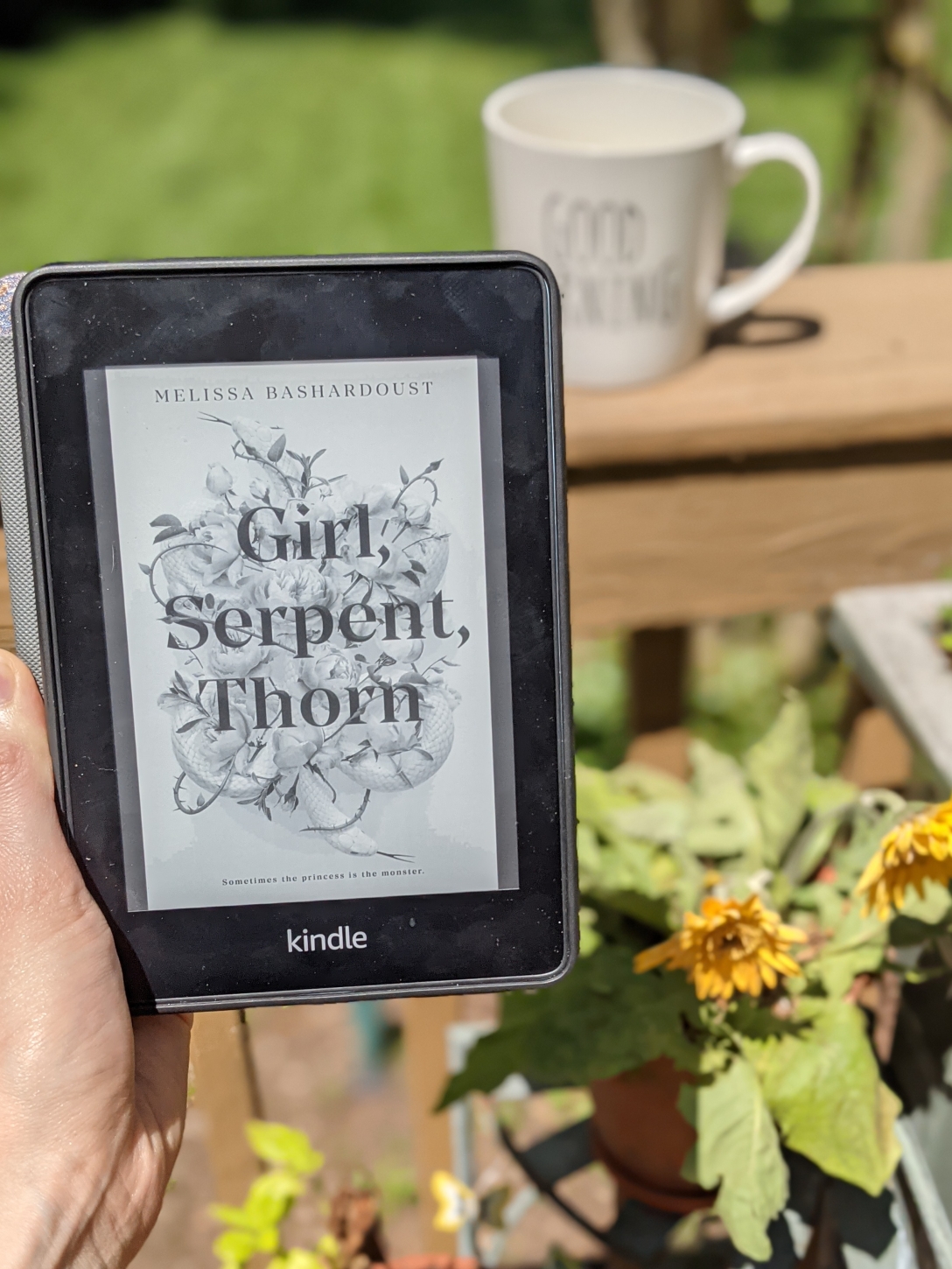 A Kindle cover of Girl, Serpent, Thorn, is held up in front of a Good Morning mug and a small pot of yellow flowers.
