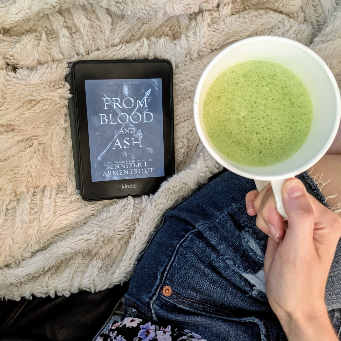 A Kindle copy of From Blood and Ash rests on a cream-colored blanket next to a white mug with a matcha latte.