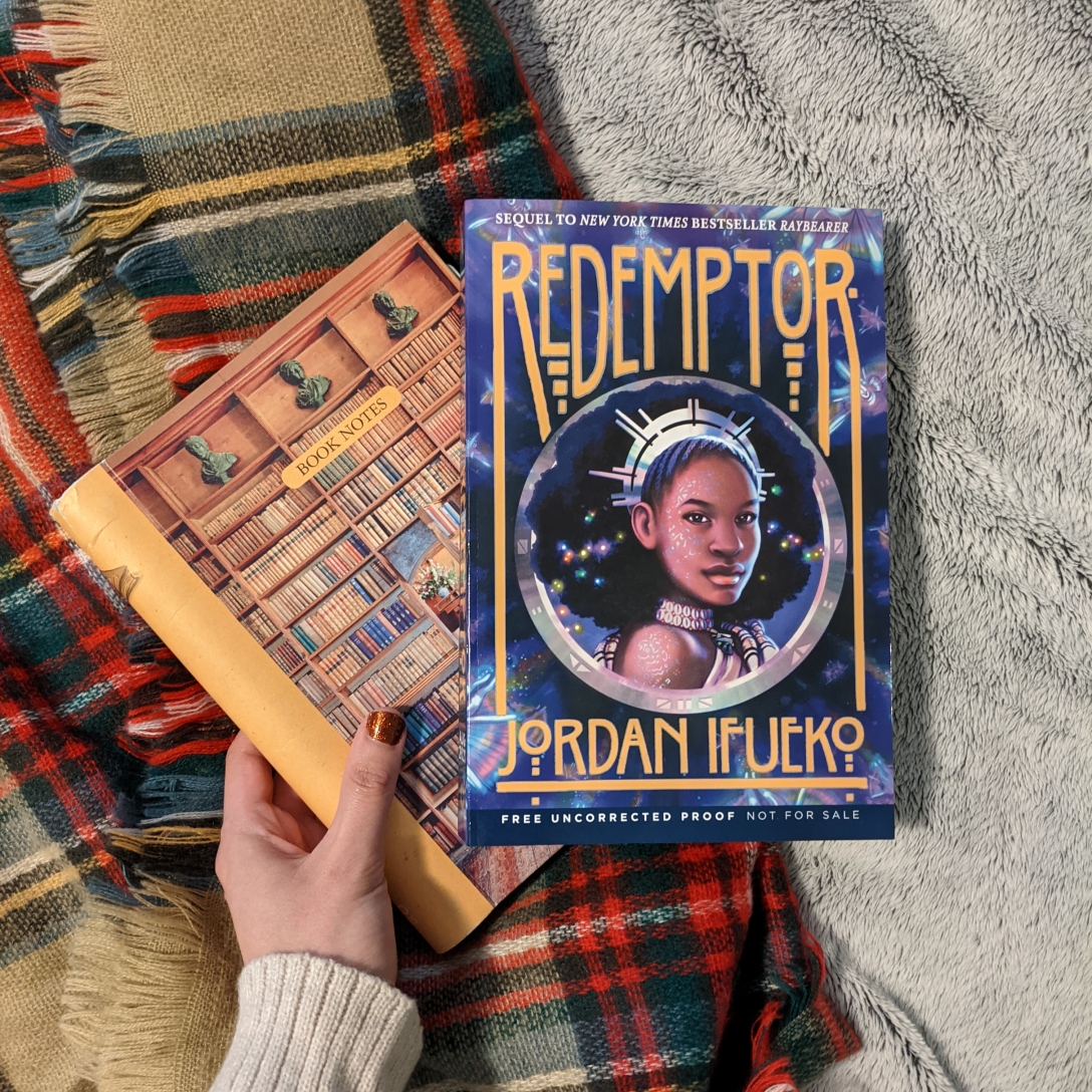 A copy of Redemptor lies on top of a book journal. Both are lying on an autumn colored scarf on a gray blanket.