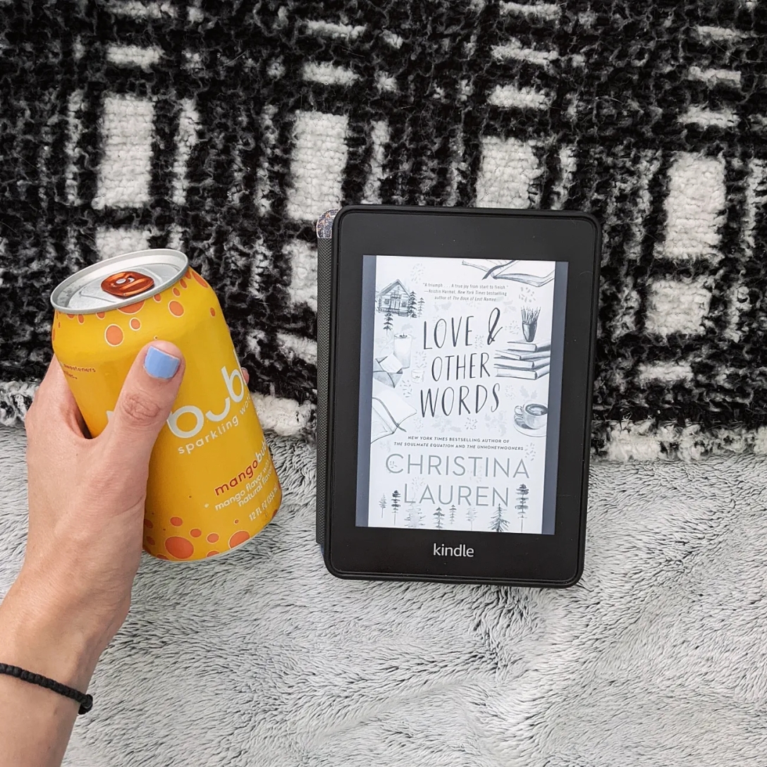 A hand holds a yellow can of mango Bubbly sparkling water next to a Kindle, which rests on both a gray blanket and a black and whtie blanket.