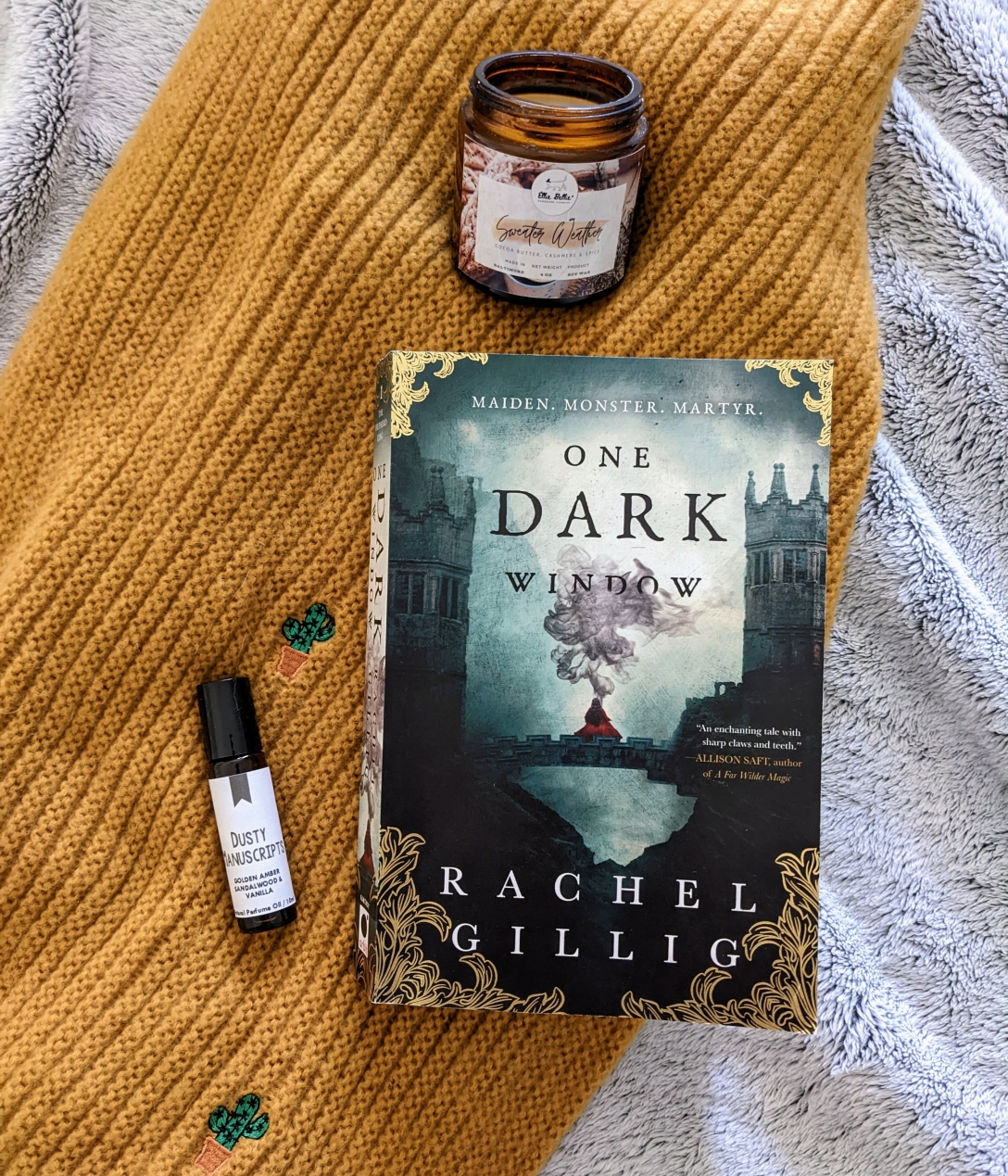 One Dark Window rests on a yellow scarf next to a cashmere candle and an oil perfume rollerball called Dusty Manuscripts.