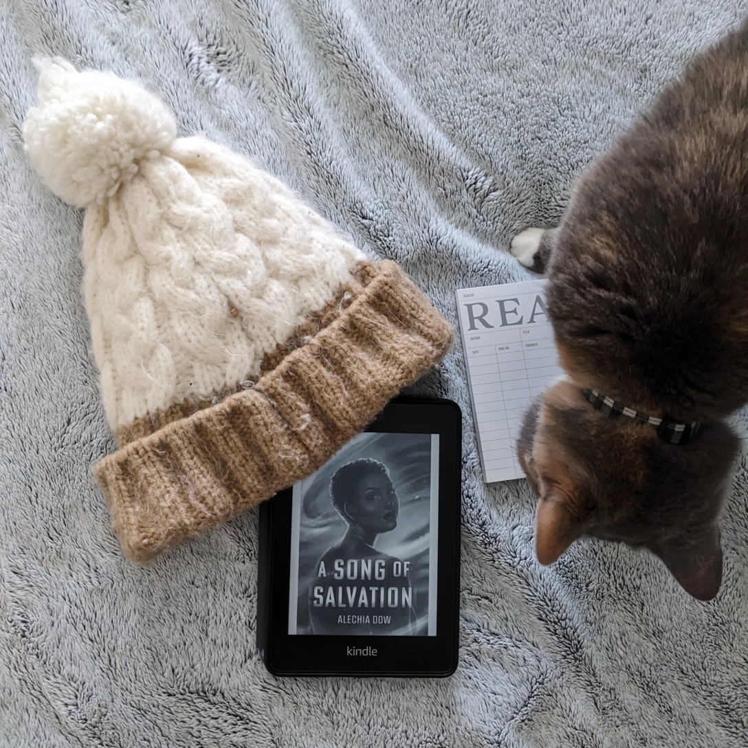 A Kindle with A Song of Salvation rests on a gray blanket next to a READ notepad, a white hat with a puff ball, and a dilute calico cat