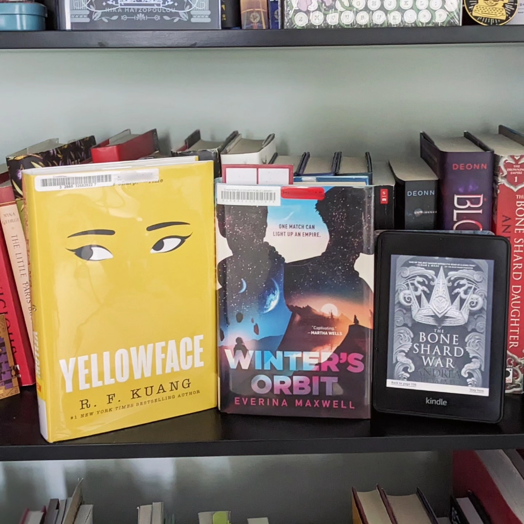 A library copy of both Yellowface and Winter's Orbit sit next to a Kindle copy of The Bone Shard War on a black bookshelf